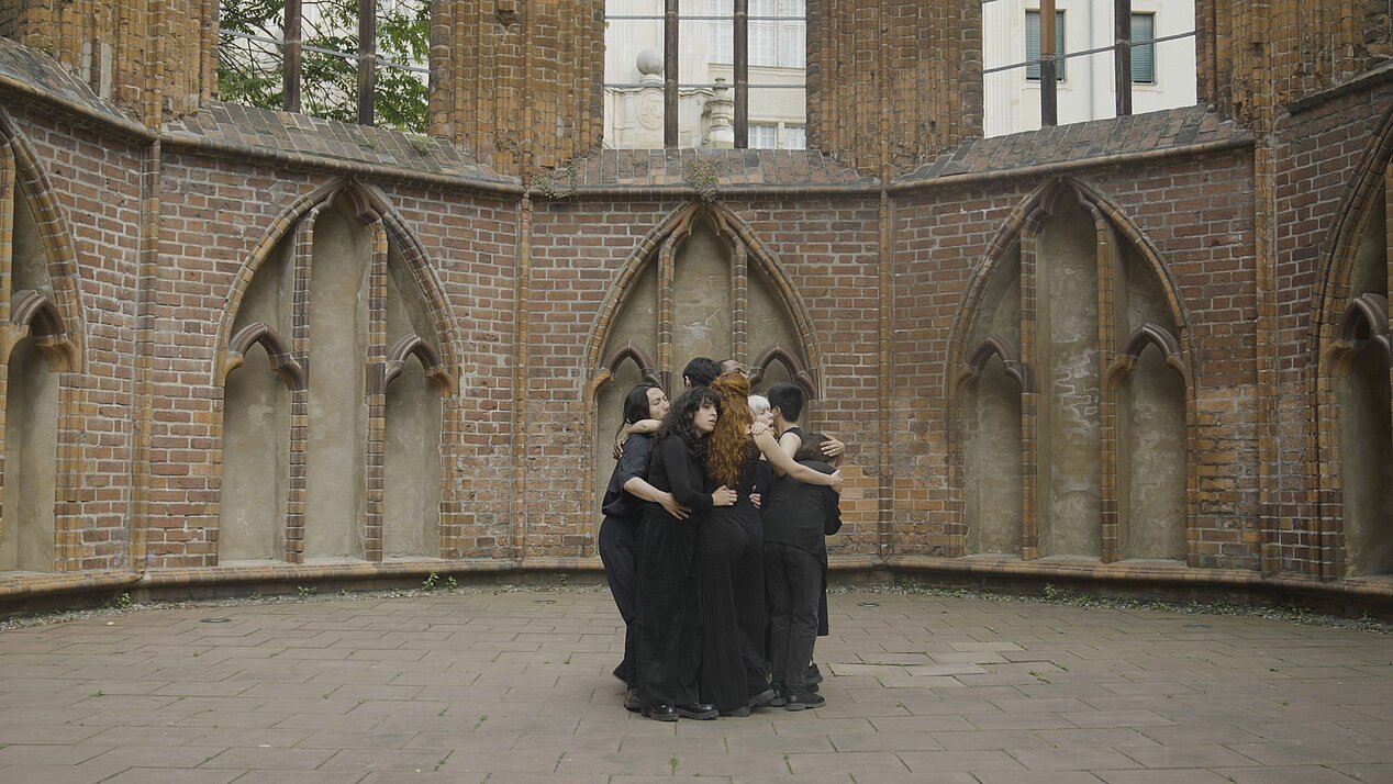 The picture is part of the exhibition Traces of Interest. It shows eight people in black clothing hugging each other tightly.