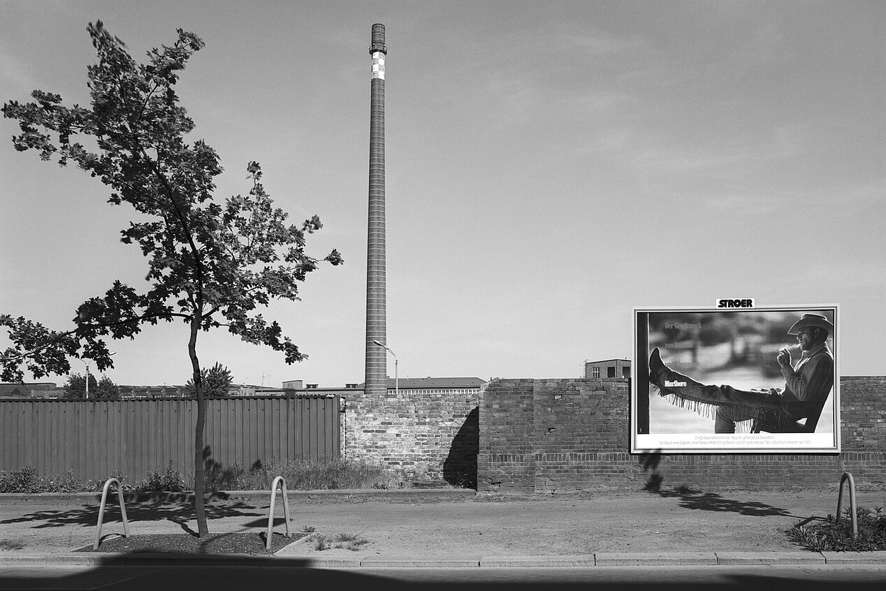 Die Fotografie ist schwarz-weiß. Links ist ein Baum, in der Mitte ein hoher Turm und rechts ein Plakat mit einem Mann. Das Plakat ist eine Marlboro Werbung.