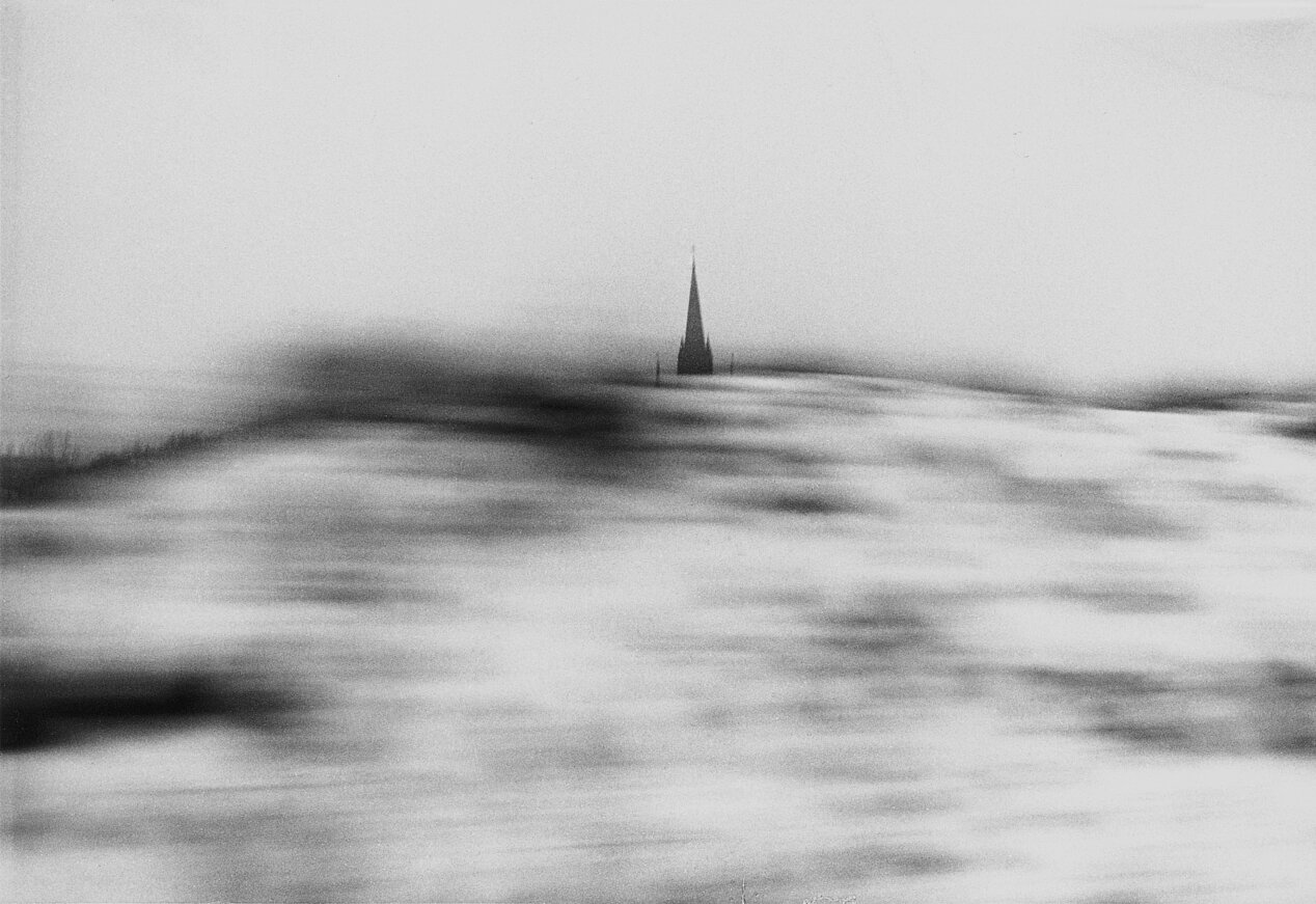 You can see a black and white picture from Helga Paris' works. You can see a building in the background, it resembles a church. There is a hill in the foreground and it is very blurred.