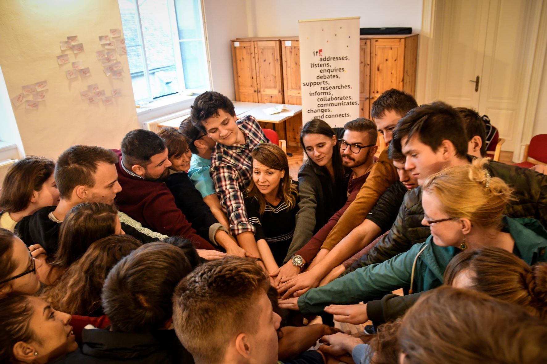 Man sieht eine Gruppe Menschen bei einem Seminar. Sie stehen im Kreis und alle halten ihre Hände in der Mitte zusammen.