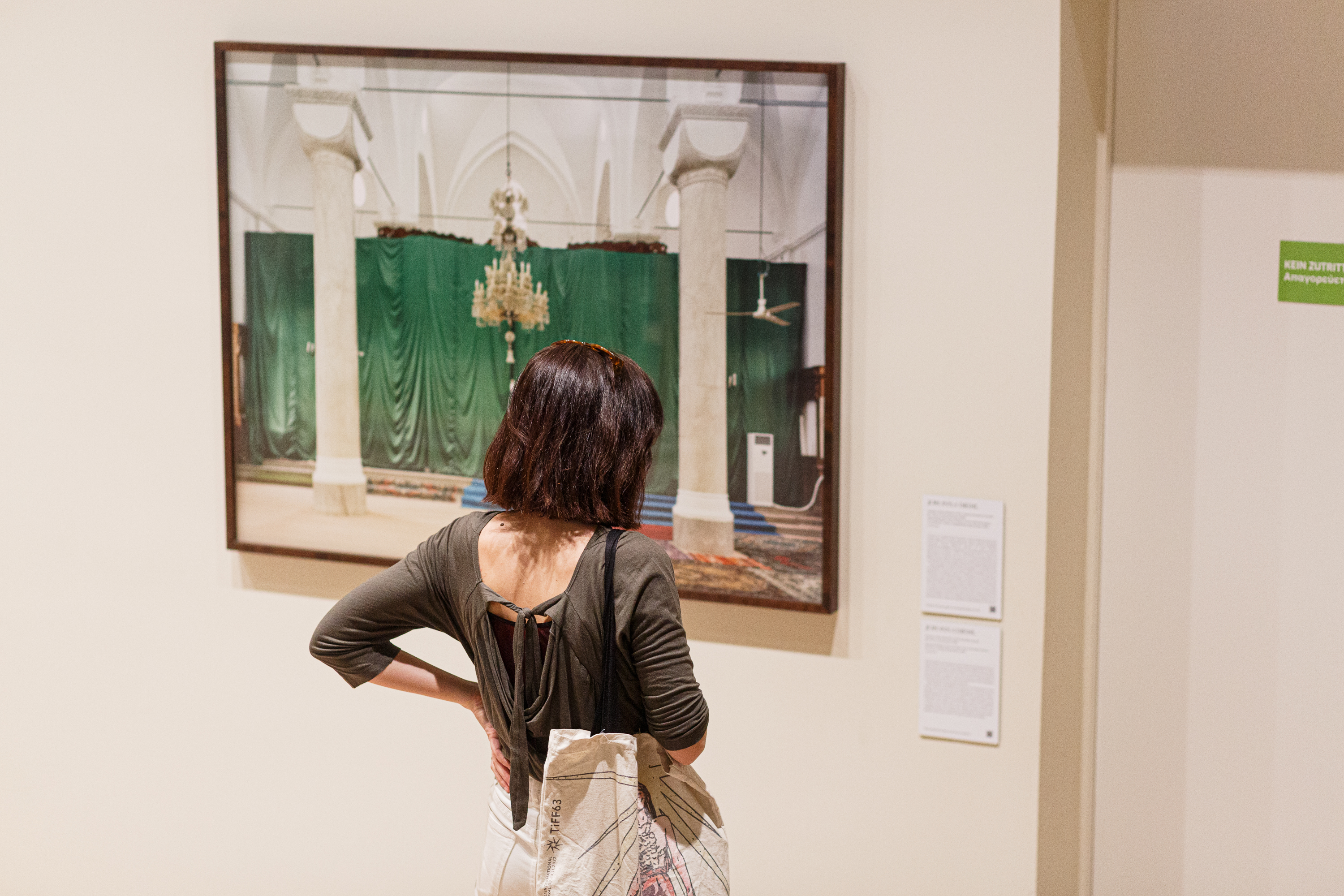 The photo shows a woman who is observing a painting on the wall. In the painting a chandelier with green curtains in the background can be seen. The photo shows the impressions from the touring exhibition "EVROVIZION" in the ifa gallery in Stuttgart, which can be visited from April 29 to June 16, 2023. © Eftychia Vlachou 