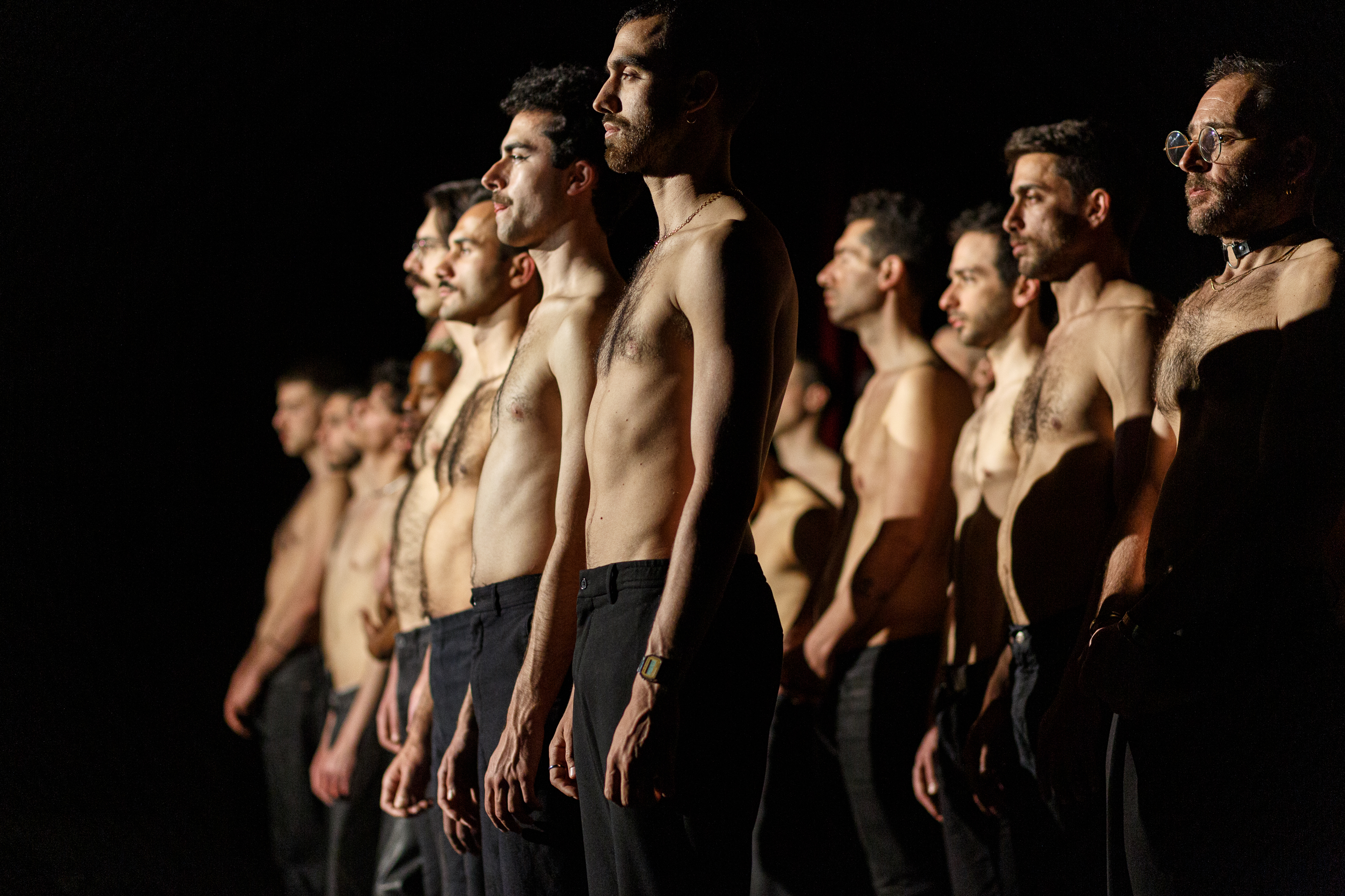 The photo shows the shirtless men in black trousers who are standing in two rows (presumably on stage). The men can be seen in the dim light while the rest of the photo is black due to the light conditions. The photo is part of the touring exhibition "EVROVIZION" in the ifa gallery in Stuttgart, which can be visited from April 29 to June 16, 2023. © Eftychia Vlachou