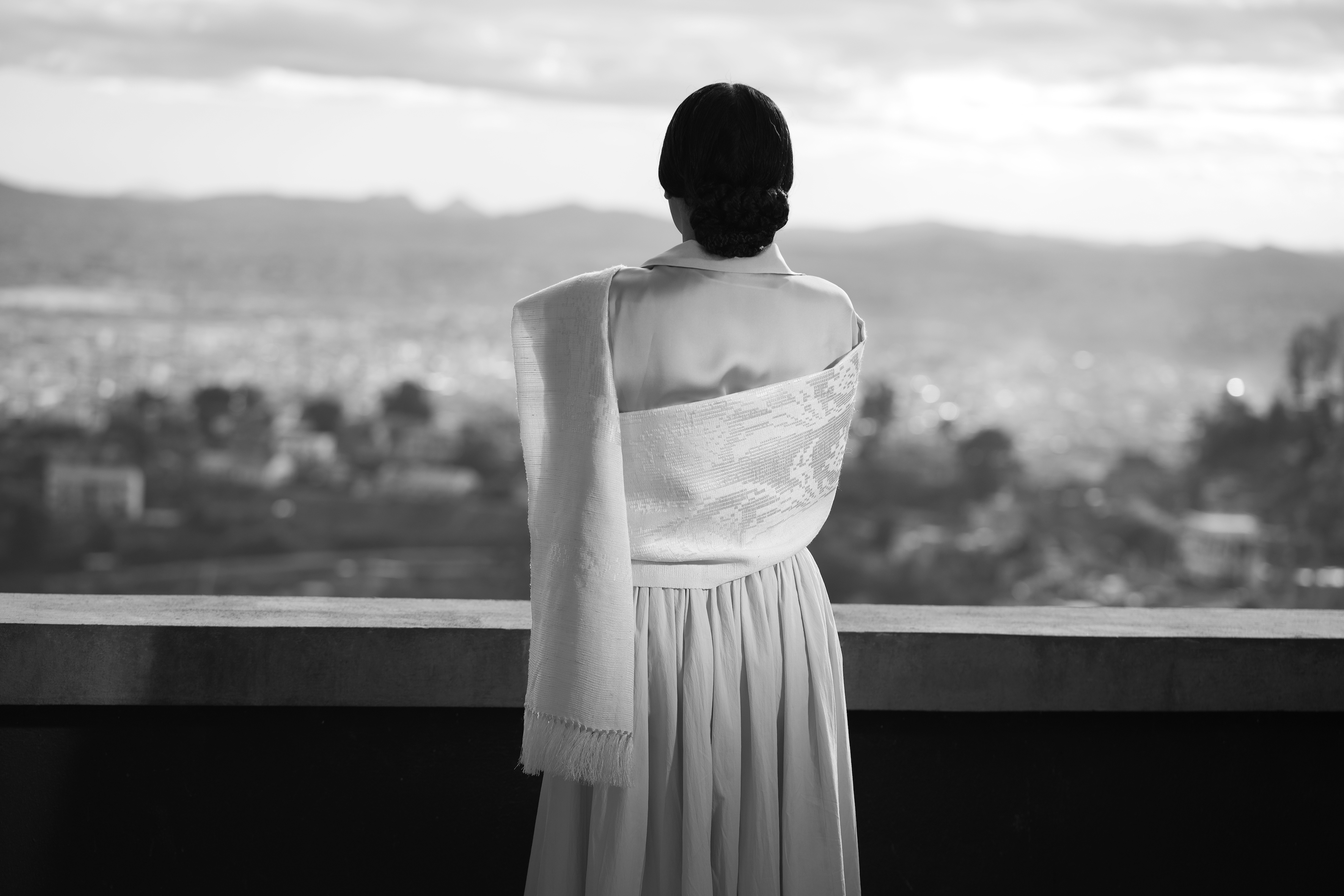 Man sieht ein film still aus Joël Andrianomearisoas PLEASE SING ME MY SONG BEFORE YOU GO. Es ist eine schwarz-weiß Aufnahme und man sieht von hinten eine Frau in einem hellen Kleid, die auf einer Art Balkon über eine Stadt blickt.
