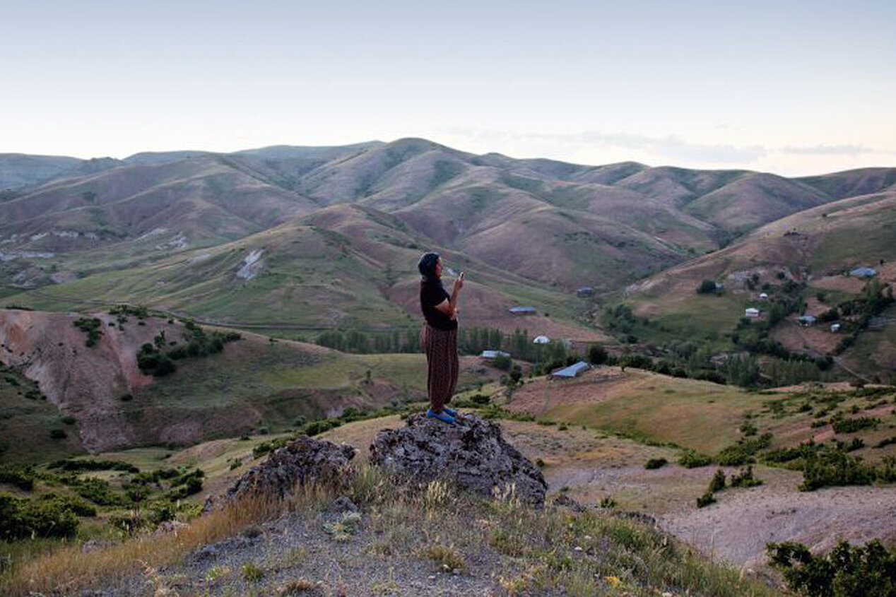 Eine Frau steht auf einem Hüge inmitten einer kargen Landschaft und hält ein Handy in ihrer Hand.