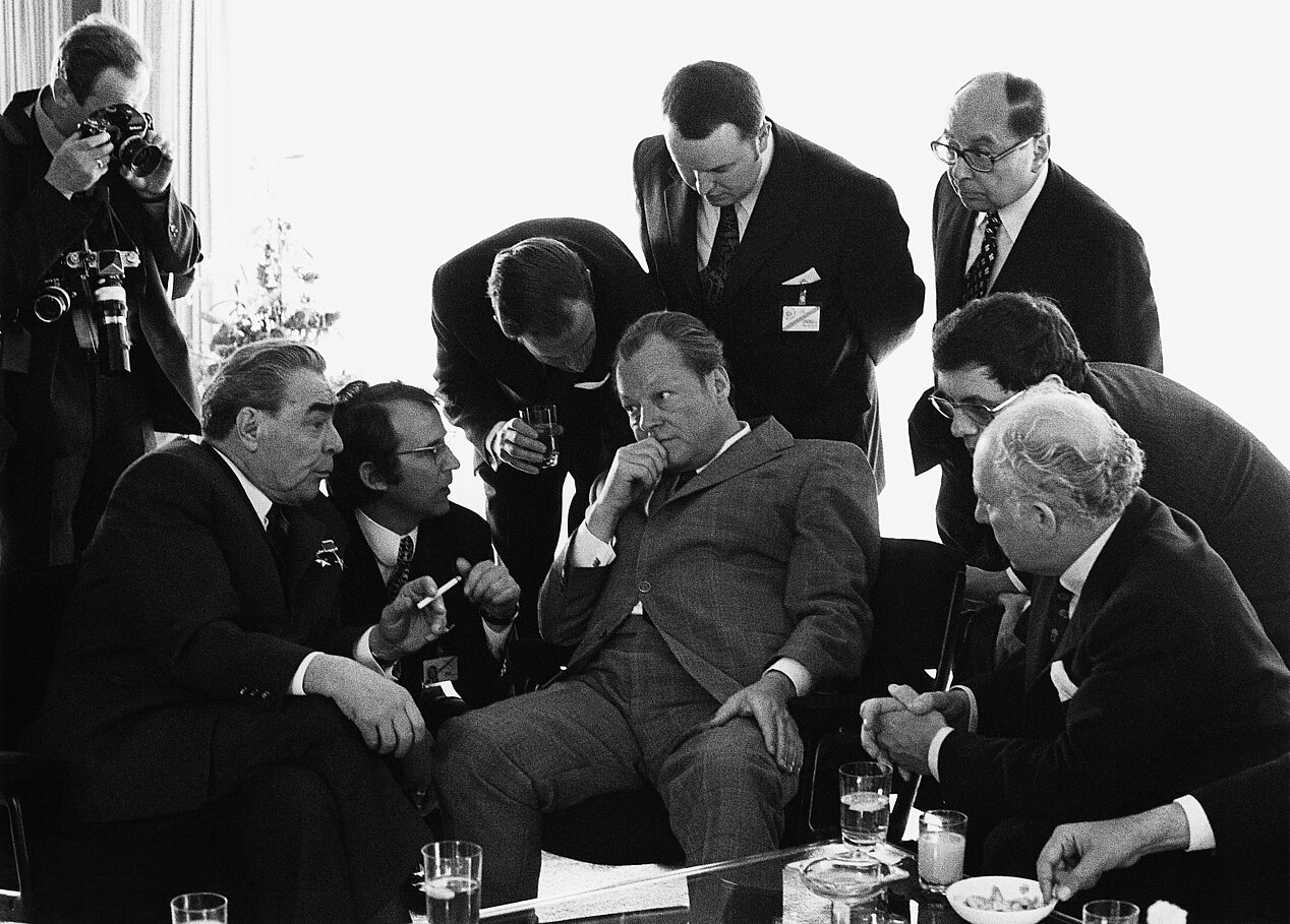 Nine men can be seen in a black and white photograph. The former German Chancellor Willy Brandt is sitting in the middle, in front of him squatting and holding a cigarette in his left hand is the Soviet politician Leonid Brezhnev. It is a photograph by Barbara Klemm, part of the ifa touring exhibition "Barbara Klemm. Light and Dark. Photographs from Germany".