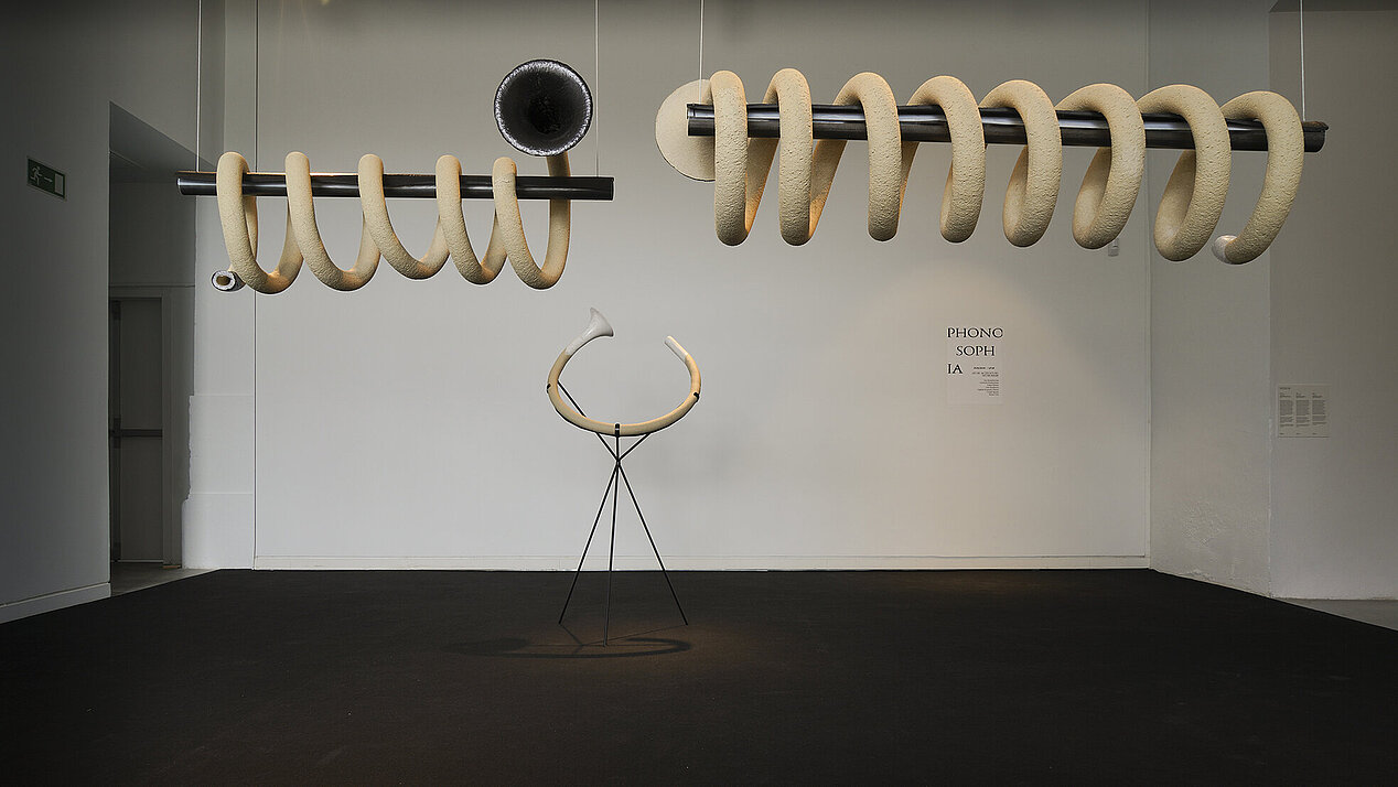 An illustration with an installation in a spiral shape and a trumpet-like ending. In the center of the image is a horn on the stand, positioned under the spiral installation. In the lower right corner on the background wall is written in black: "Phonosophia". It is the opening of the exhibition "Camila Sposati – Breath Pieces" in ifa-Galerie Stuttgart, which can be visited on 12. May 2023. © Sascha Fronczek