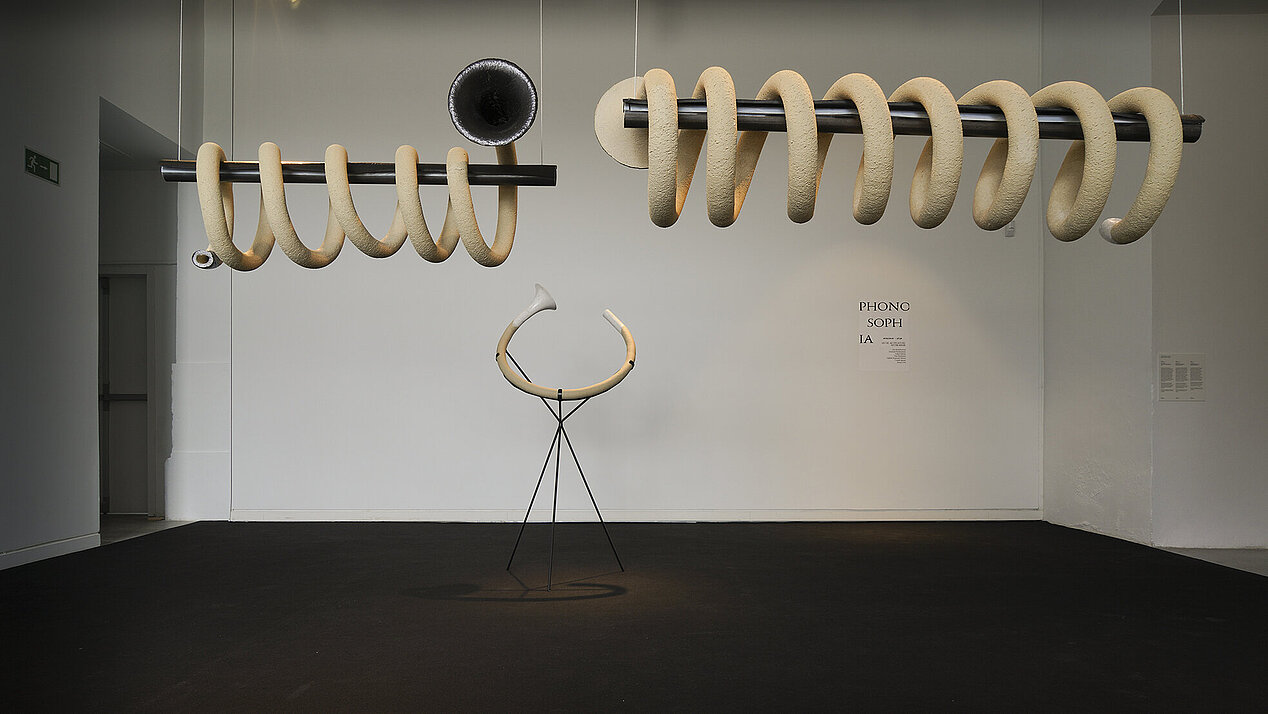 Eine Installation in Spiralform und trompetenähnlichem Ende. In der Mitte des Bildes befindet sich ein Horn auf dem Gestell, das unter der spiralförmigen Installation positioniert ist. In der rechten unteren Ecke an der Wand im Hintergrund steht in schwarzer Schrift: "Phonosophia". Es handelt sich um die Ausstellungseröffnung "Camila Sposati. Atem-Stücke" in der ifa-Galerie Stuttgart, die am 12. Mai 2023 zu besuchen ist. © Sascha Fronczek