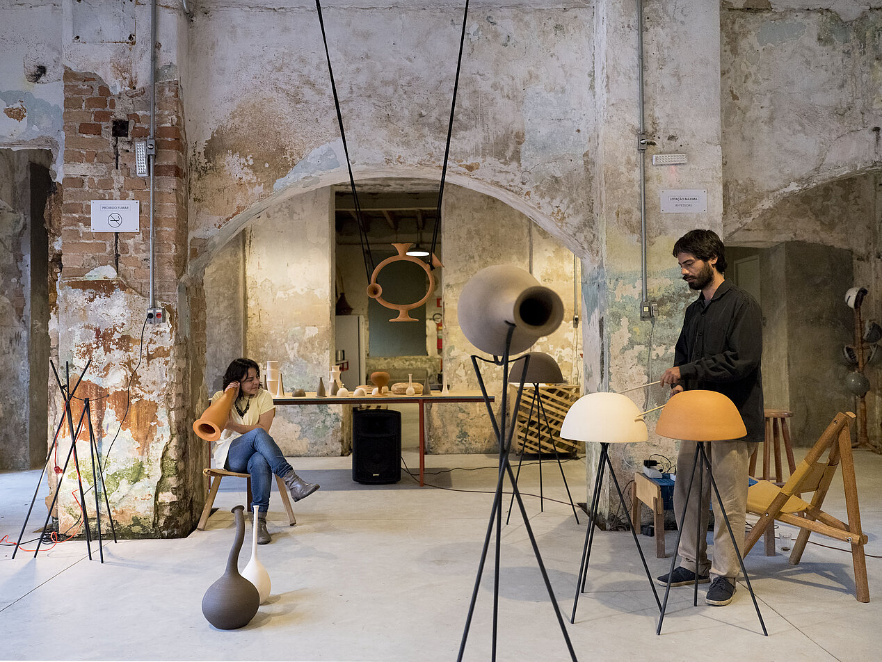 The photo shows a view of the exhibition Phonosophia by the Brazilian artist Camila Sposati. In a room with high ceilings and partially white-plastered brick walls, two people can be seen, a woman is sitting on a chair on the left, a man with a beard is standing on the right. The woman is pressing her ear to some kind of clay instrument, the man seems to be playing with two wooden sticks on one of the two upturned clay bowls. There are more of these clay sculptures standing and hanging in the room. 