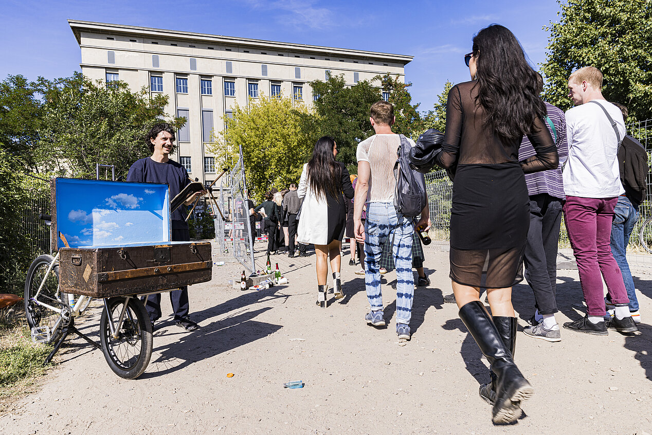 You can see Adrien Missaka's art object MOTUS, which is mounted on a bicycle. The artist and the bicycle are standing in front of the Berghain club. Many people are walking towards the club. The sun is shining.