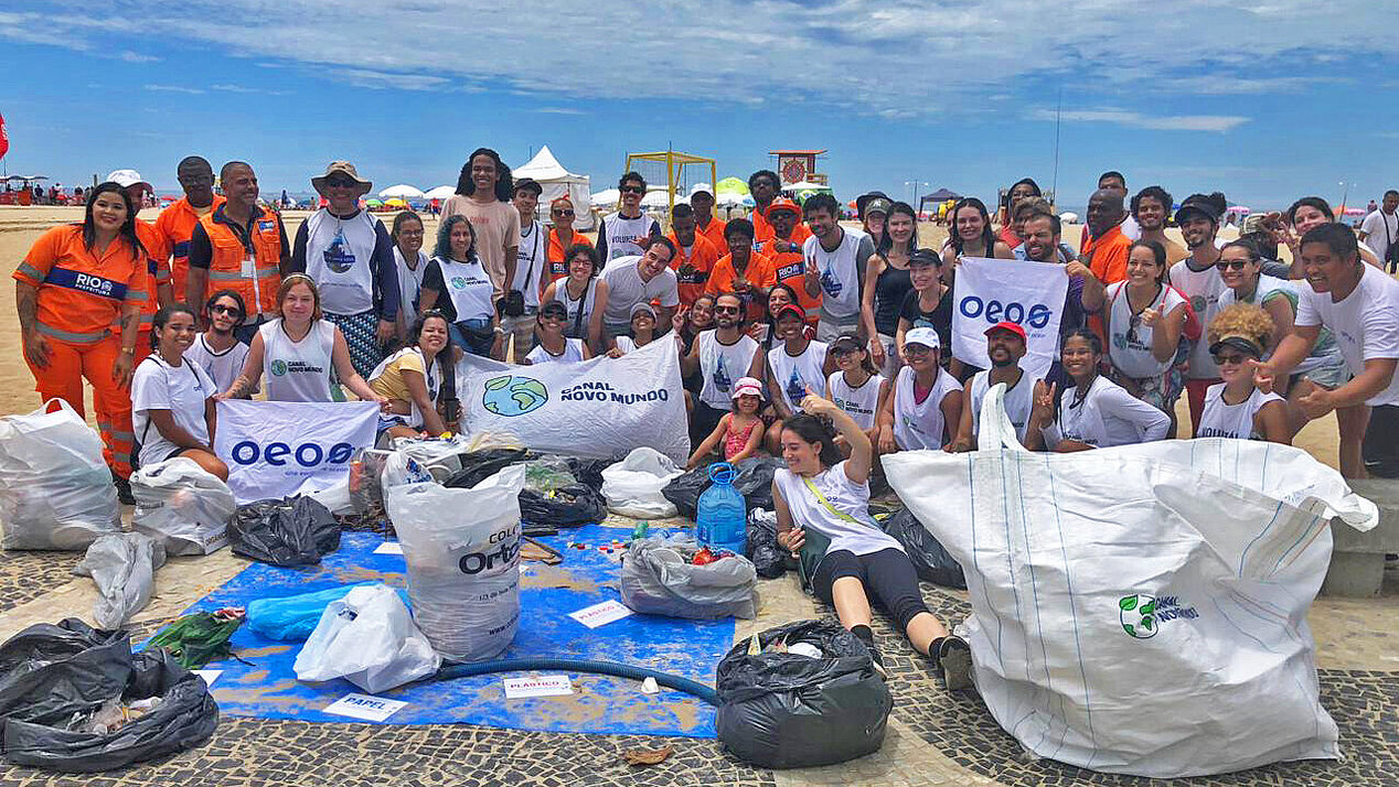 The photo shows the environmental activists in Brazil, members of Canal Novo Mundo and One Earth One Ocean. In front of the activists posing on a beach are full garbage bags. The photo is from the series of environmental actions organized by Thaine Maciel in Brazil.