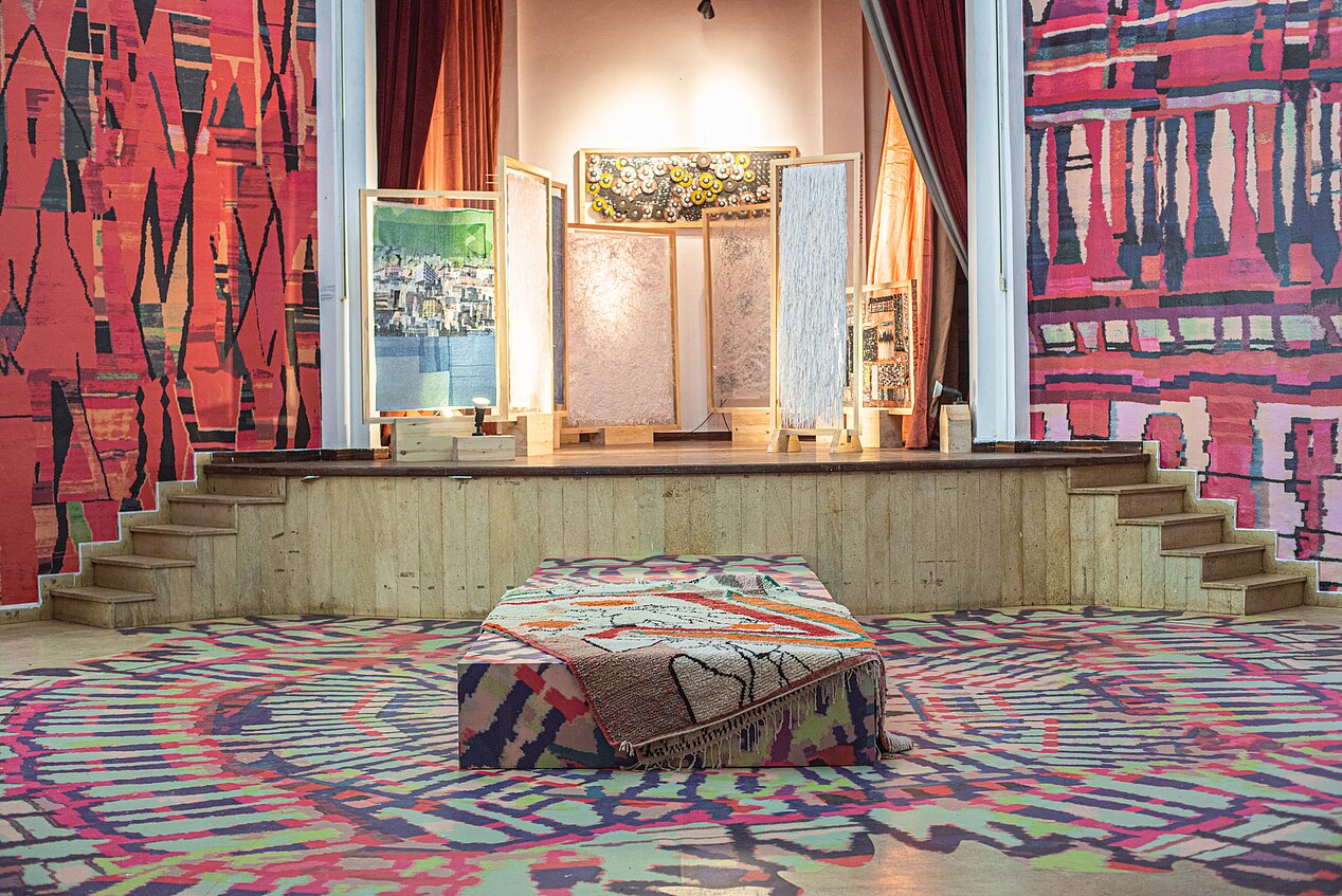 Das Bild zeigt eine Ausstellungsansicht der ifa-Galerie Berlin aus der Ausstellung School of Casablanca. Man blickt in einen Raum und im Hintergrund sieht man eine Bühne, mit zwei kleinen Treppen rechts und links. Auf der Bühne sind Bilder aufgestellt. Der Raum ist nicht bestuhlt, sondern mit Teppichen ausgelegt. An den Wänden sind auch Teppiche. In der Mitte des Raumes ist ein etwas erhöhtes Podest, ebenfalls mit Teppichen bedeckt.