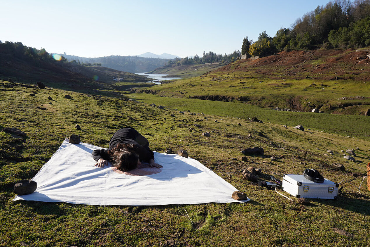 Eine Person in schwarzer Kleidung liegt auf einem weißen Tuch inmitten grüner Landschaft.