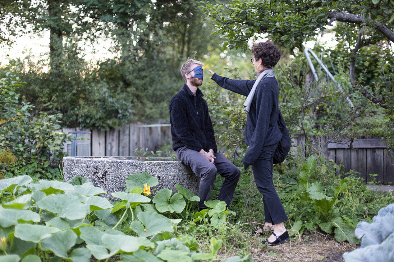 Sitzender Mann in einem Garten mit verbundenen Augen, vor ihm steht eine Frau, die ihn an der Stirn berührt. Szene aus Listening Session #1