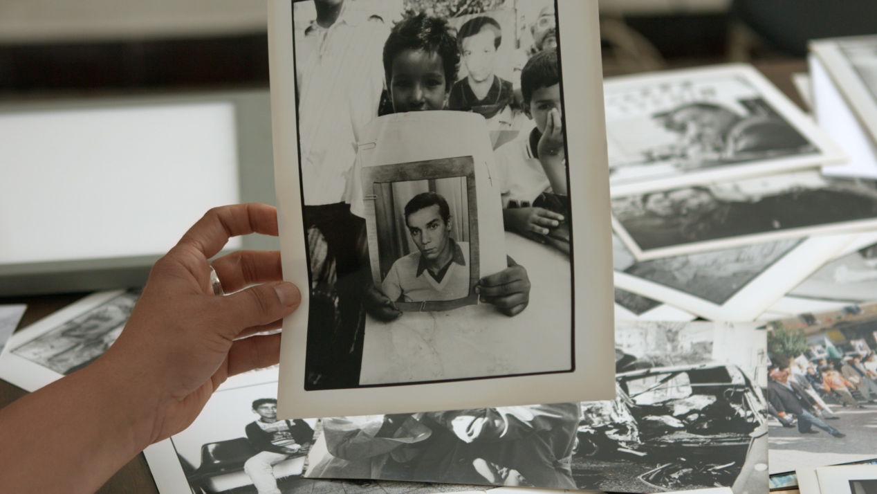 The picture shows a black and white photo. The photo shows a child holding another photo in his hands and presenting it to the viewers. The photo within a photo shows a man with a serious look. In the background one can see more black and white photos on the table. The picture is part of the film screening "Le Roman Algérien", which will be shown as part of the exhibition "The Canticle oft he Birds" from May 13 to July 30 at the ifa Gallery in Berlin.