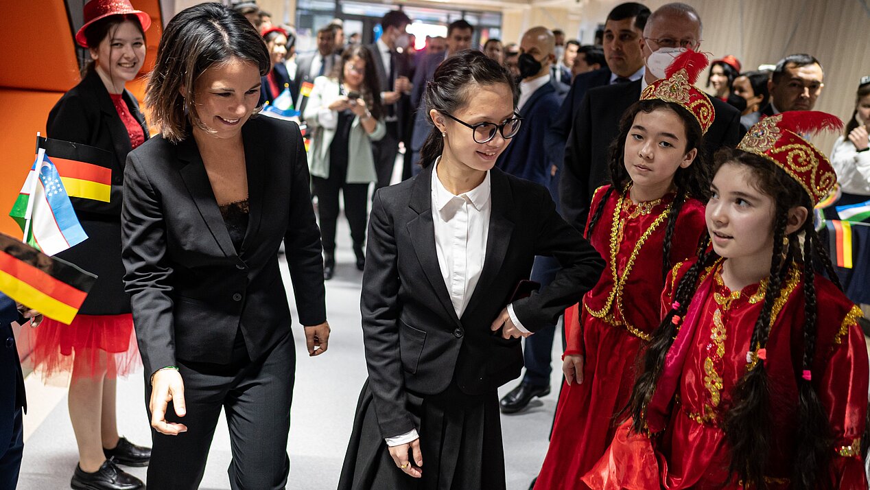 Foreign Minister Annalena Baerbock wears a black trouser suit. She smiles as she walks through a group of children in a corridor. She is accompanied by a young woman in a costume with glasses. Both look at two little girls to their right. The girls are wearing red velvet costumes decorated with golden ornaments. In the background are people holding small flags of Germany and Uzbekistan.