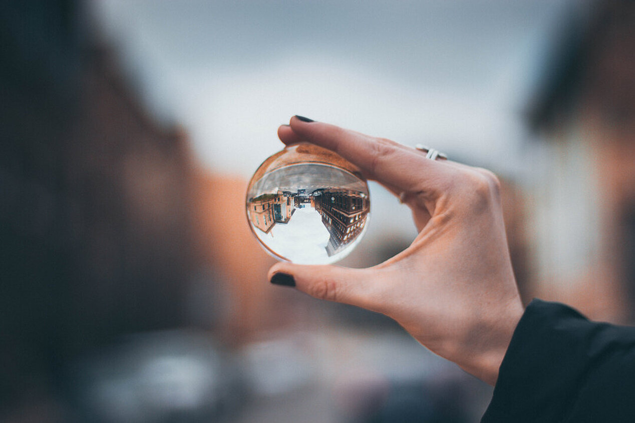 Eine Hand hält eine Glaskugel, in der eine Stadtansicht gespiegelt ist.
