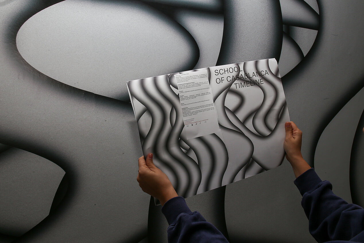 In the photo, two hands are holding a piece of paper in the air. The paper reads School of Casablanca and a small text. The background of the paper and the picture in general are black and white wavy lines.