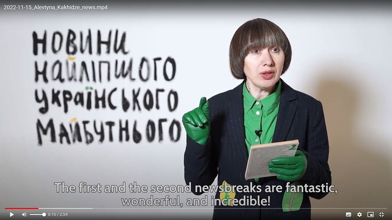 The photo shows the artist Alevtina Kakhidze, wearing green leather gloves, holding a notebook and gesturing while explaining. A heading in Cyrillic script can be seen in the background. The subtitle shows the following text: "The first and the second newsbreaks are fantastic, wonderful and incredible!" The photo represents a sequence from the video record and it is a part of the workshop "All Times News from Stuttgart and Other Cities", which takes place in the ifa gallery in Stuttgart. The workshop is related to the exhibition "Art and Life in Times of War".