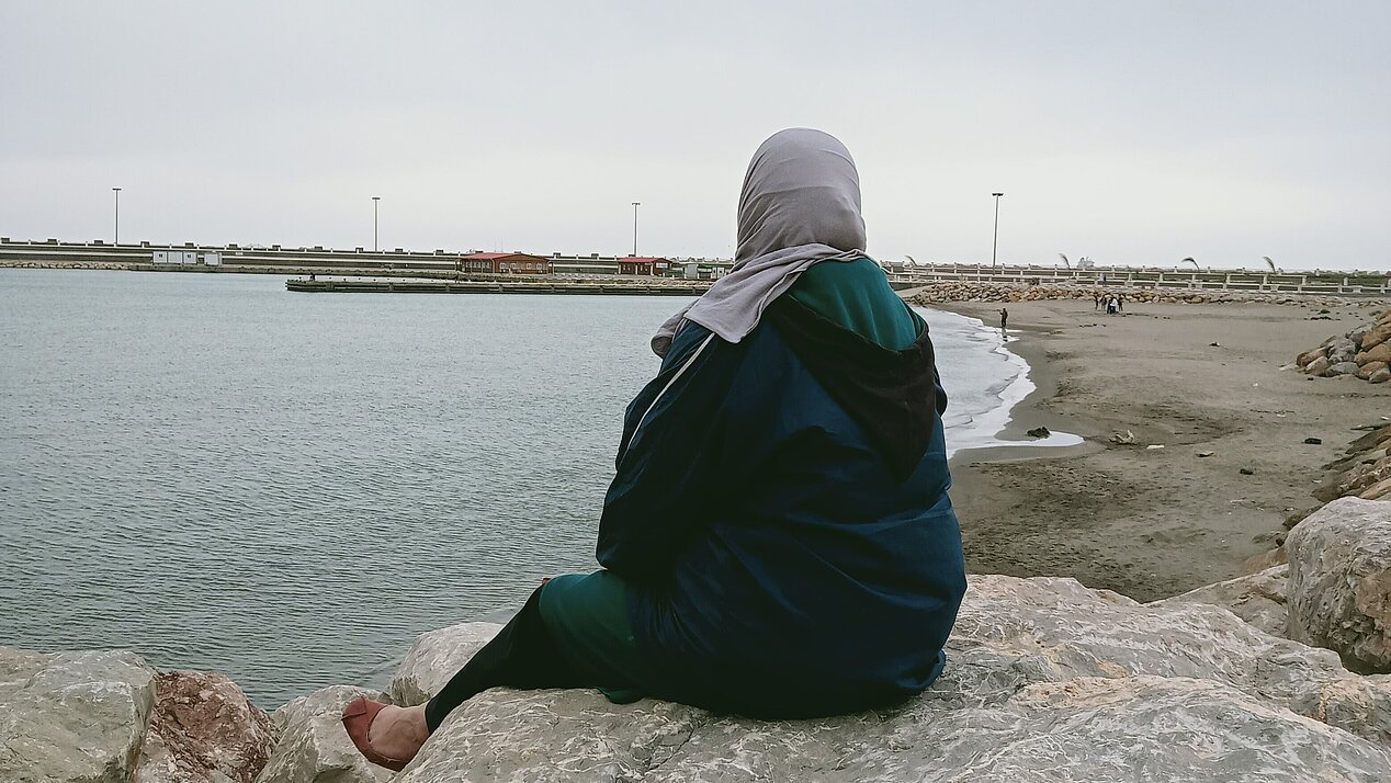 A woman with scarf sitting on a stone at a beach, watching into the far ocean. Photograph: Zineb Bettayeb