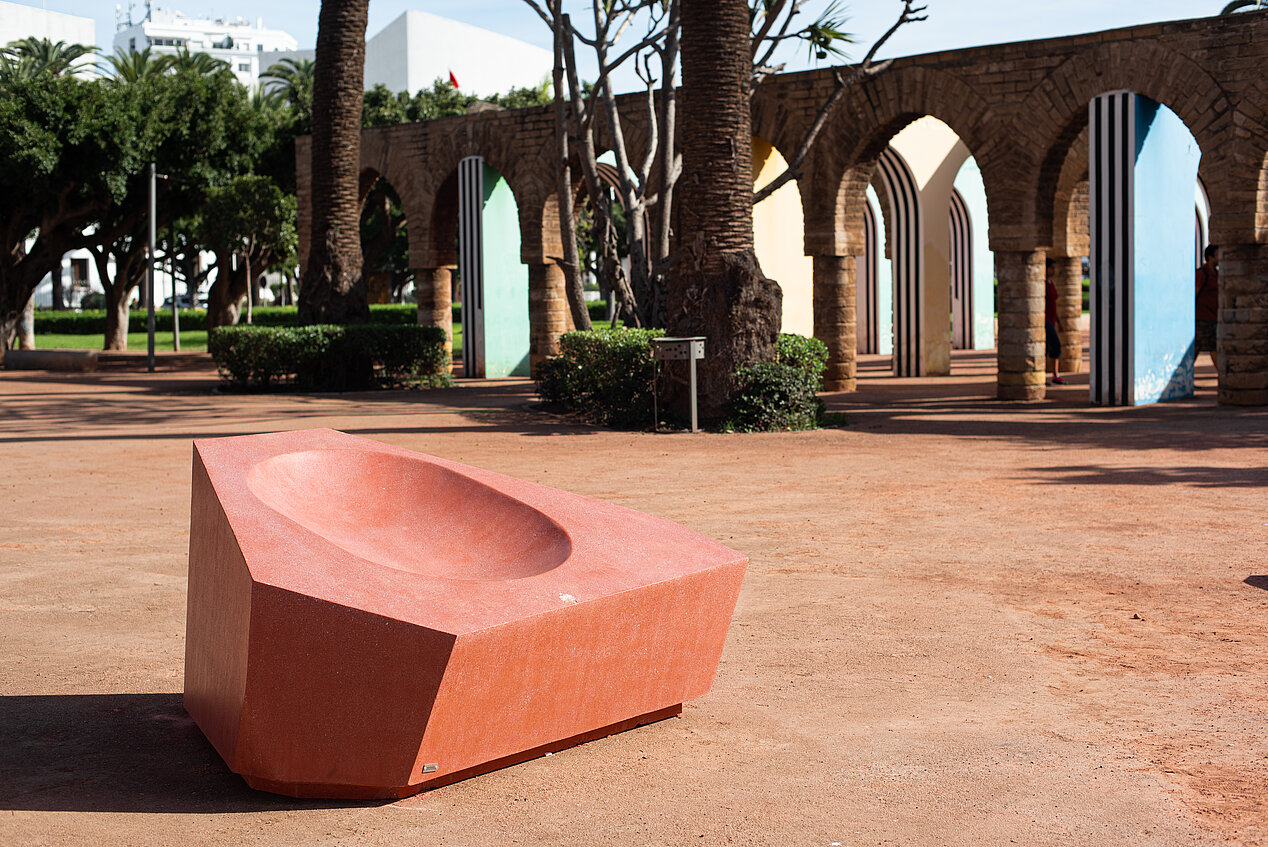 Das Bild zeigt eine Ausstellungsansicht der School of Casablanca. Es ist im Freien aufgenommen. Im Hintergrund sind Palmen und Torbögen zu sehen. Der Boden ist rötlich-sandfarben und im Vordergrund ist eine rostrote Skulptur.