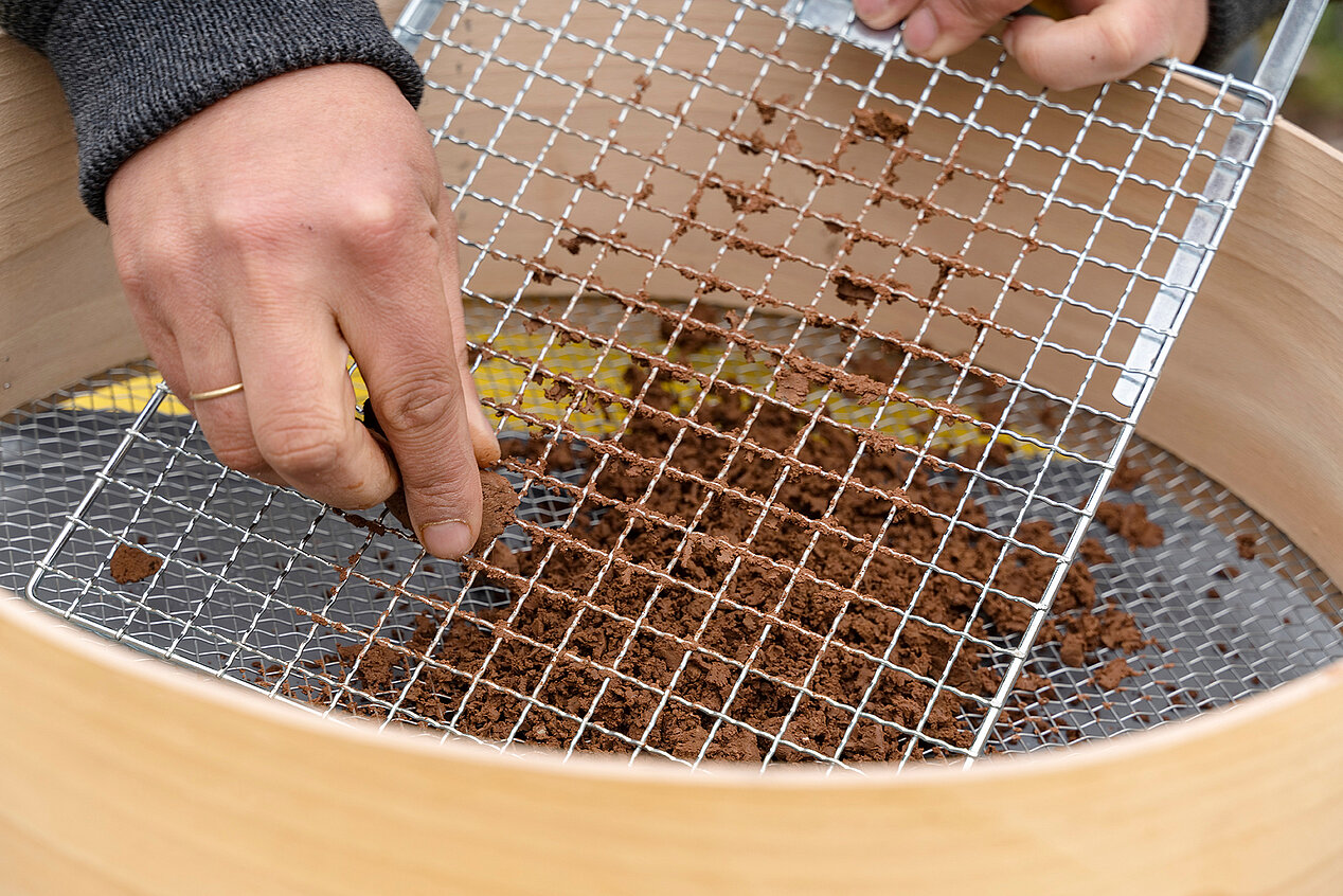 A large sieve can be seen in the picture. In the center of the picture is a hand rubbing clay with the help of a net. It is the cover to a family workshop linked to the exhibition Camila Sposati – Breath Pieces at the ifa Gallery in Stuttgart.