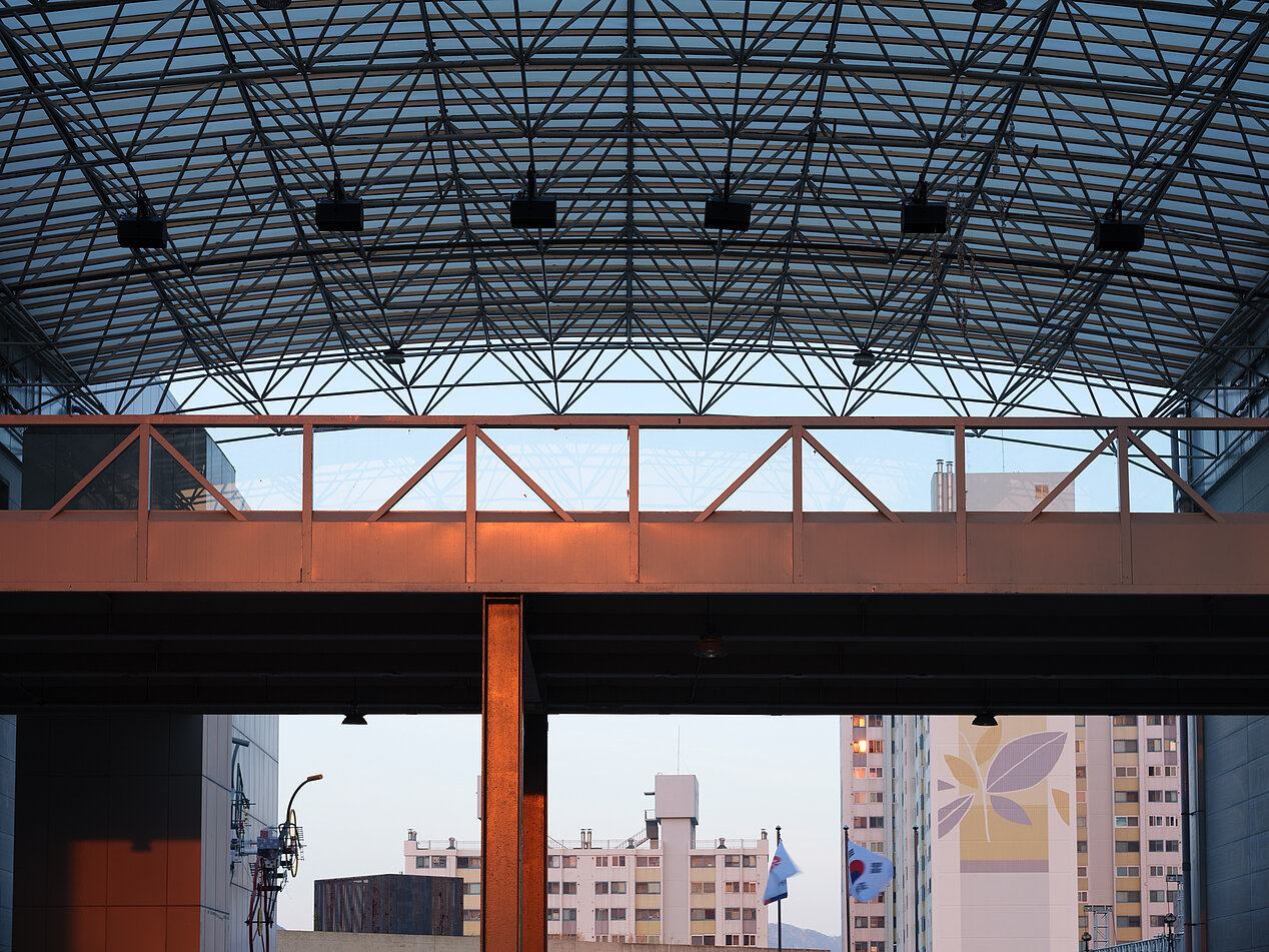 The photo shows a construction with a mechanical roof under which a red bridge can be seen. In the background there are apartment buildings, with the Korean flag flying under the bridge. The photo is a part of the 14th Gwangju Biennial taking place from April 7th to July 9th in Gwangju, Republic of Korea. © Pan Daijing