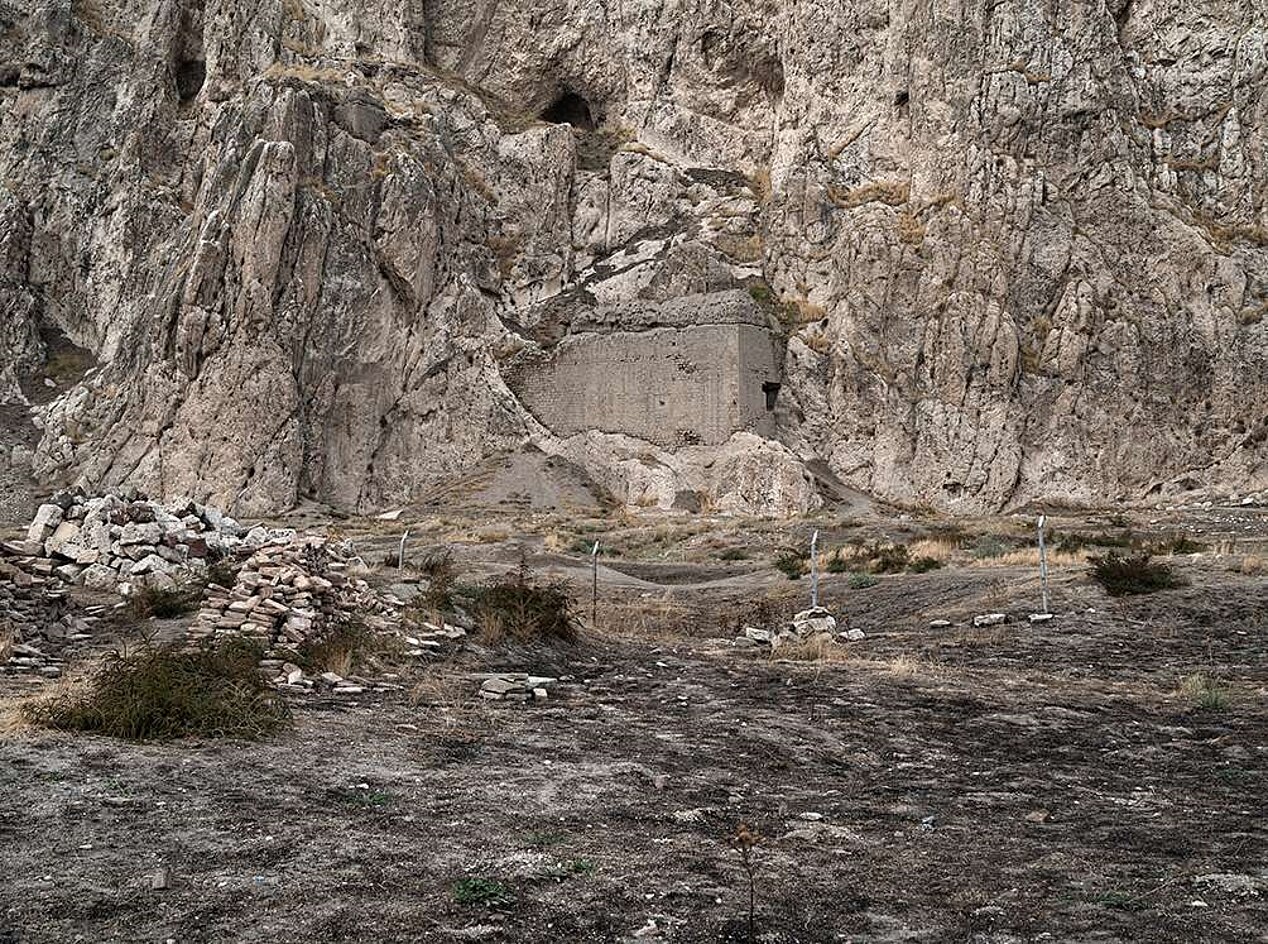 A bleak landscape in front of a steep rock face can be seen. A few green bushes and shrubs grow out of the barren ground, a wire fence can be faintly made out and a concrete structure is built into the rock face. This is a work by the artist Andréas Lang, whose exhibition BROKEN MEMORIES was supported by ifa's exhibition funding.