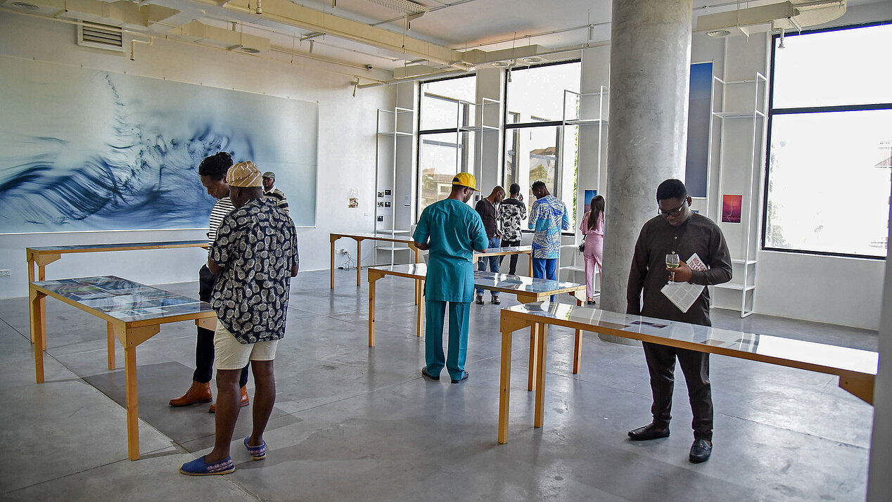 Eröffnung der Tourneeausstellung "Fragile" von Wolfgang Tillmans in Lagos: in einem hellen Raum stehen Menschen verteilt und blicken auf Tische vor sich. Die Menschen sind schwarz. Auf den Tischen liegen verschiedene Objekte aus der Fotografie-Ausstellung von Wolfgang Tillmans, man erkennt nicht genau was. An der Wand ist eine große Fotografie einer blauen Welle. © Goethe-Institut Nigeria