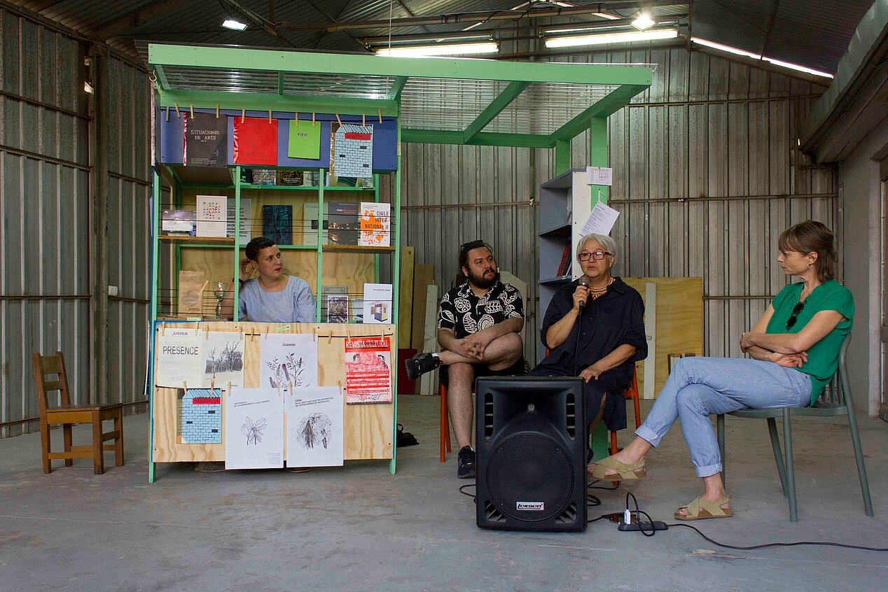 Four people are seated in a hall. The people are sitting in or next to a wooden kiosk. In the front, there is a large loudspeaker and one of the four people is currently talking into a microphone. It is the opening speech of the ifa-funded exhibition "EL KIOSCO" and book publication of CHILE INTERNATIONAL II. The people are Valeria Fahrenkrog, Diego Parra, Ana María Saavedra, Eva-Christina Meier (from left to right). Photo: Ash Aravena, 2023