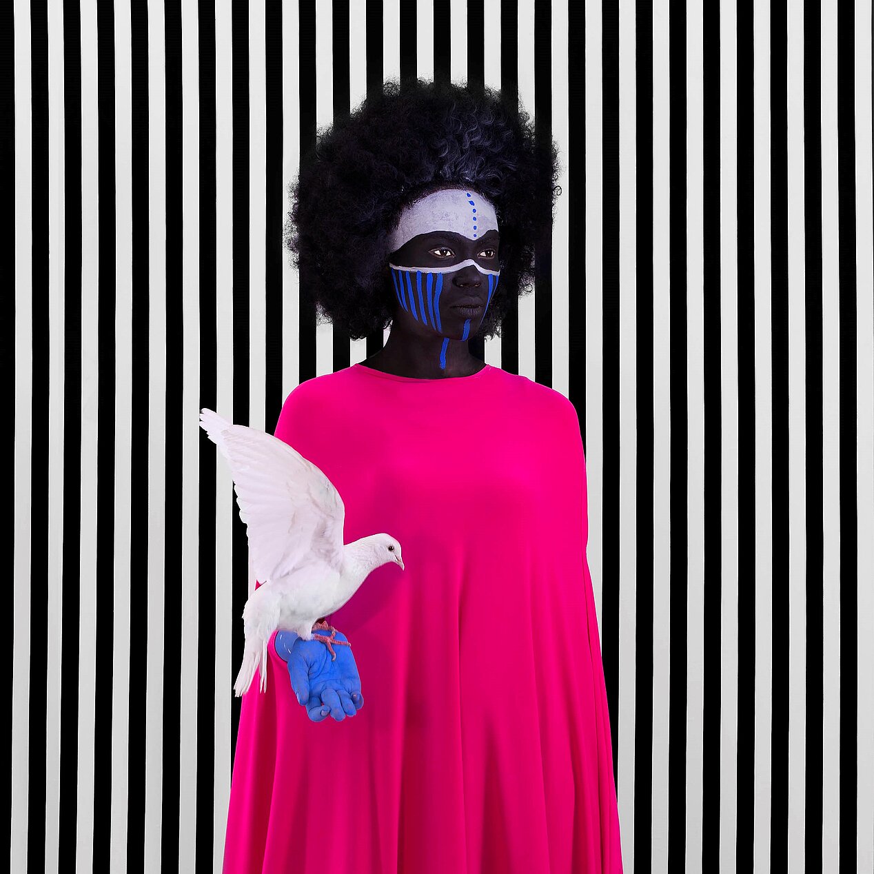 The picture shows the artist Aïda Muluneh in a pink dress. Her face is painted white and blue, her right hand is painted blue. A dove is sitting on her right hand. The background has black and white stripes.